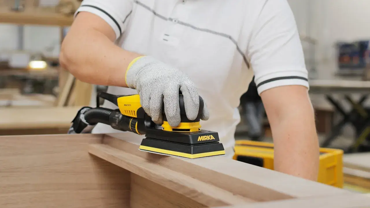 Un artisan utilise une ponceuse Mirka pour comment poncer un meuble avec des moulures. La ponceuse est jaune et noire, et l'artisan porte des gants de protection.