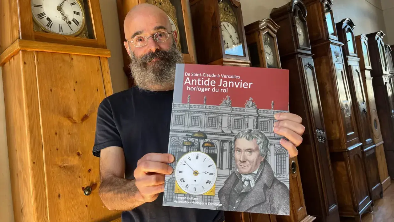Un homme barbu présente un livre sur Antide Janvier, horloger du roi, devant une rangée de grandes horloges anciennes.