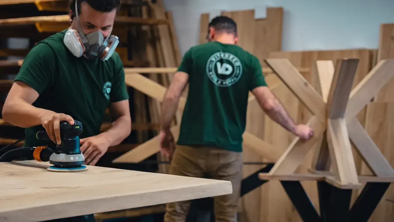 Un artisan masqué utilise une ponceuse pour poncer une table en bois. Un autre artisan travaille sur un pied de table en bois en arrière-plan.