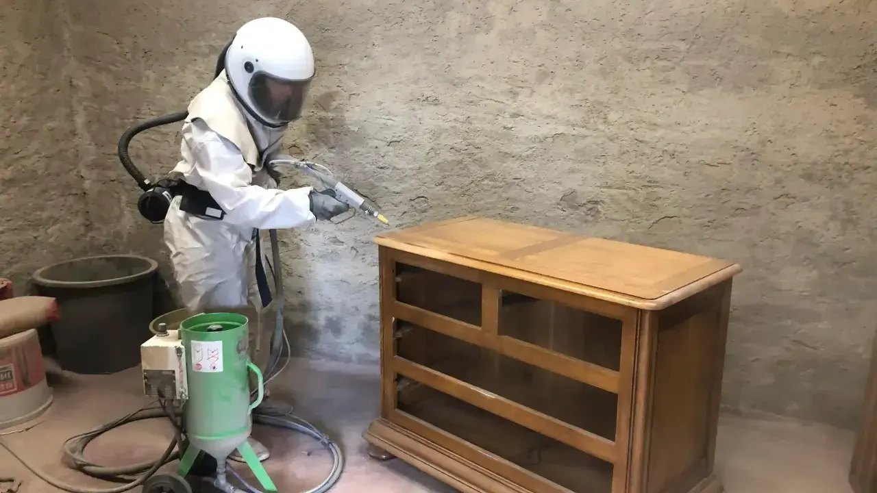 Personne en combinaison intégrale et casque effectuant un décapage bois sur une commode ancienne avec une machine de sablage.