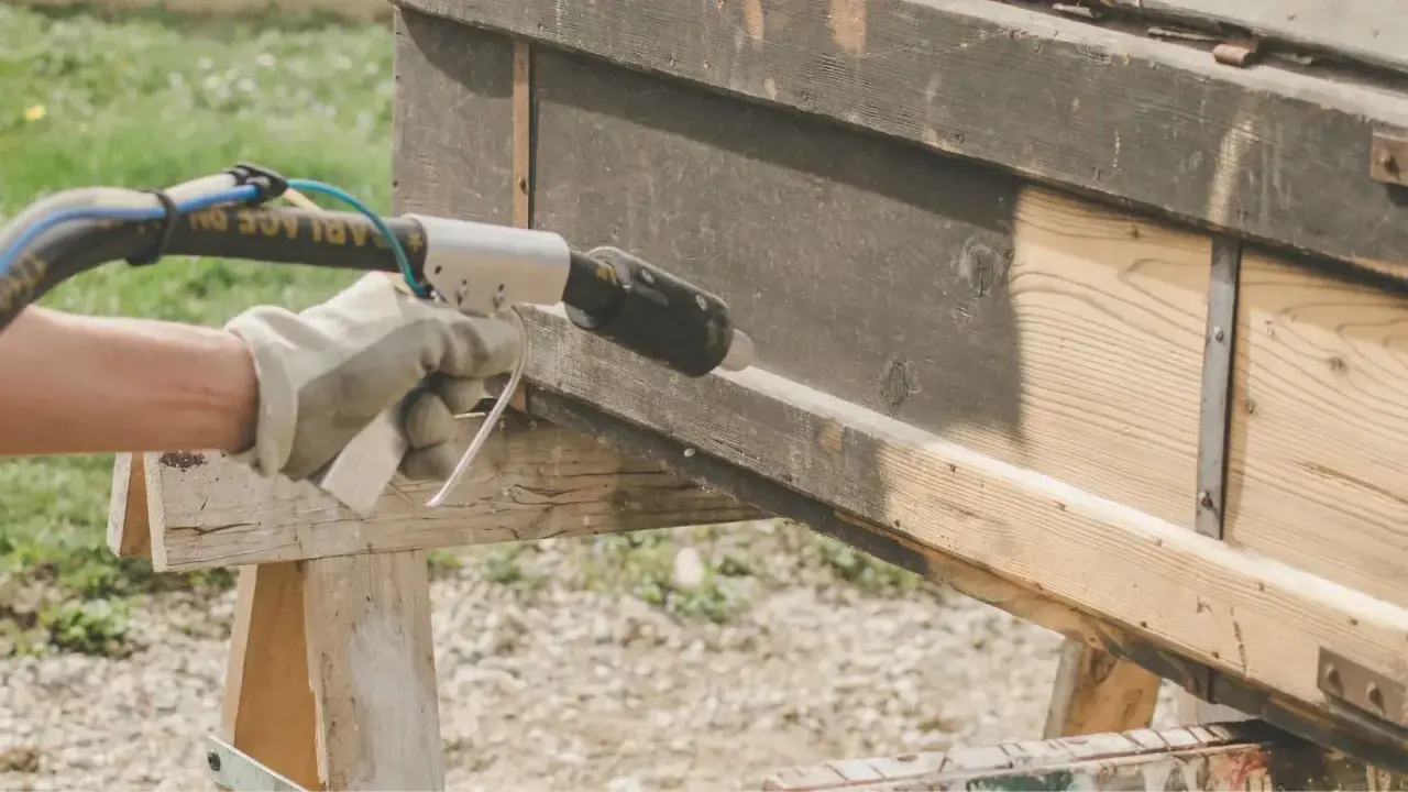 Main tenant en décapeur thermique pour enlever peinture sur bois. Le bois brut apparaît sous la couche sombre.