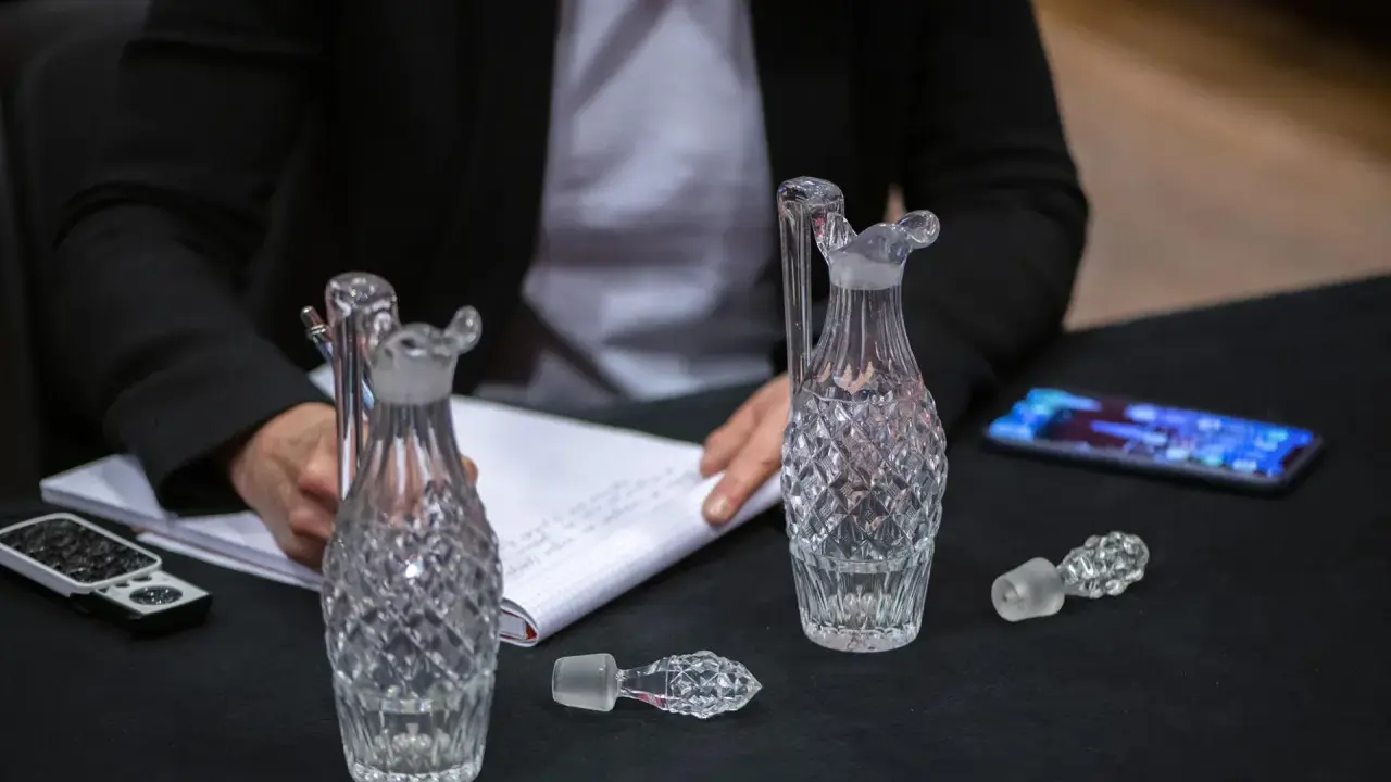 Une personne écrit sur un carnet, entourée de flacons en cristal. Une folle enchère se prépare pour ces objets précieux.