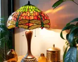 Lampe tiffany origine, avec abat-jour en vitrail coloré orné de libellules, posée sur une table en bois près de plantes vertes.