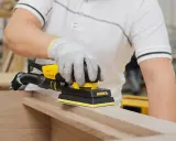 Un artisan utilise une ponceuse Mirka pour comment poncer un meuble avec des moulures. La ponceuse est jaune et noire, et l'artisan porte des gants de protection.