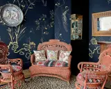 Salon au style napoléon 3, avec des fauteuils et un canapé en rotin corail, sur un tapis léopard, devant un papier peint bleu nuit orné de motifs dorés.