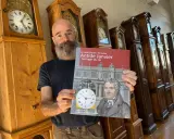 Un homme barbu présente un livre sur Antide Janvier, horloger du roi, devant une rangée de grandes horloges anciennes.