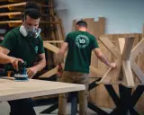 Un artisan masqué utilise une ponceuse pour poncer une table en bois. Un autre artisan travaille sur un pied de table en bois en arrière-plan.