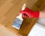 Application d'huile de lin sur un bois clair. La main gantée de rouge applique le produit pour protéger le bois, loin des insectes.