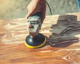 Main d'artisan utilisant une ponceuse pour enlever lasure sans poncer le bois. Le grain du bois est bien visible.