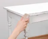 Main tenant, comment décaper un meuble peint. Une main utilise du papier de verre pour poncer le bord d'une table blanche écaillée.