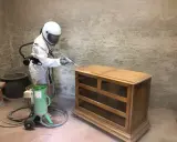 Personne en combinaison intégrale et casque effectuant un décapage bois sur une commode ancienne avec une machine de sablage.