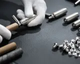 Mains gantées assemblant une ferrure de meuble. Des pièces métalliques et des vis sont disposées sur une surface sombre.