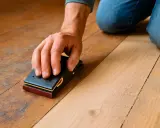 Main d'un artisan ponçant un plancher de bois avec un bloc abrasif, démontrant qu'il est possible de poncer sans ponceuse.