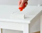 Main blanche d'un meuble laqué, un rouleau de peinture blanc est appliqué.