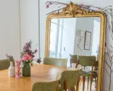 Un grand miroir ancien doré, orné de branches fleuries, reflète une salle à manger avec des chaises vertes. Quelle couleur peindre un miroir ancien ? L'or reste un classique intemporel.