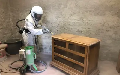 Personne en combinaison intégrale et casque effectuant un décapage bois sur une commode ancienne avec une machine de sablage.