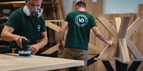 Un artisan masqué utilise une ponceuse pour poncer une table en bois. Un autre artisan travaille sur un pied de table en bois en arrière-plan.