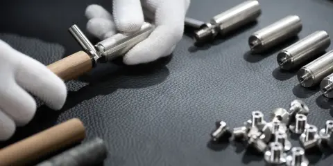 Mains gantées assemblant une ferrure de meuble. Des pièces métalliques et des vis sont disposées sur une surface sombre.
