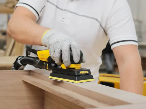 Un artisan utilise une ponceuse Mirka pour comment poncer un meuble avec des moulures. La ponceuse est jaune et noire, et l'artisan porte des gants de protection.