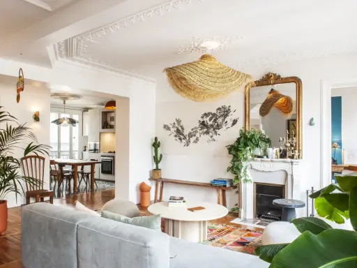 Salon au style classique revisité, avec un canapé gris, un tapis coloré, une cheminée ornée et des plantes luxuriantes.