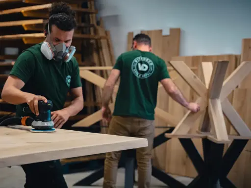 Un artisan masqué utilise une ponceuse pour poncer une table en bois. Un autre artisan travaille sur un pied de table en bois en arrière-plan.