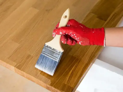 Application d'huile de lin sur un bois clair. La main gantée de rouge applique le produit pour protéger le bois, loin des insectes.