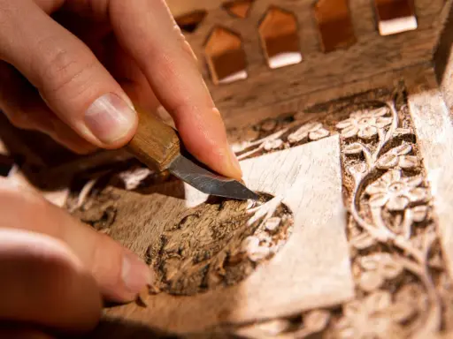 Main d'artisan sculptant un motif floral sur un meuble en bois, étape clé pour savoir comment nettoyer un meuble en marqueterie.