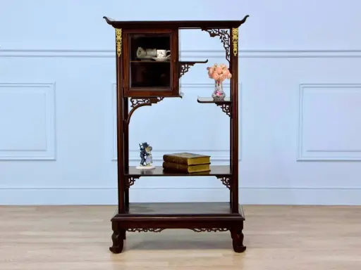 Étagère en bois sombre ornée de motifs dorés, rappelant le style de Gabriel Viardot. Elle présente des tasses dans une vitrine, des livres et des fleurs.
