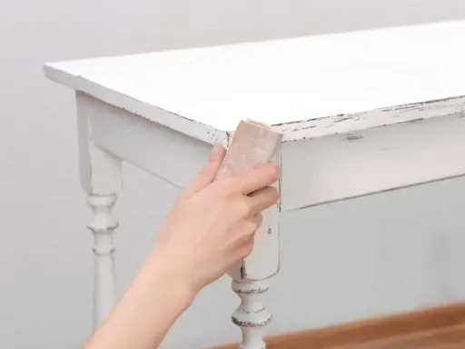 Main tenant, comment décaper un meuble peint. Une main utilise du papier de verre pour poncer le bord d'une table blanche écaillée.