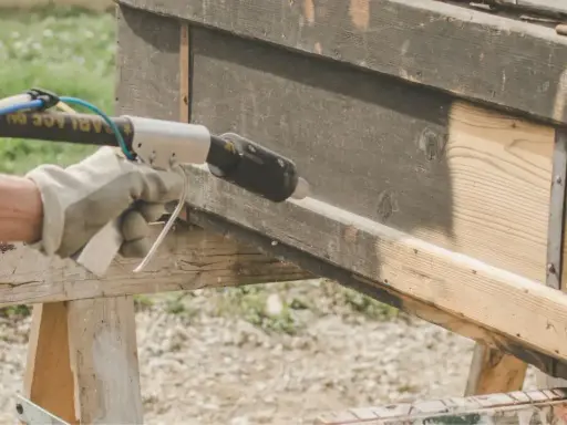 Main tenant en décapeur thermique pour enlever peinture sur bois. Le bois brut apparaît sous la couche sombre.