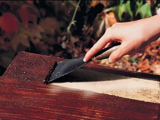 Main d'une personne utilisant un grattoir pour décaper peinture sur bois avec bicarbonate.
