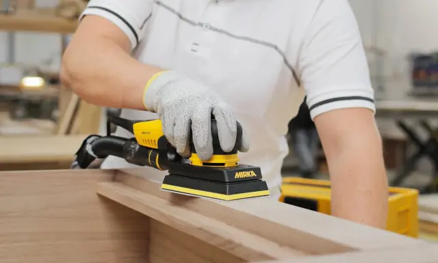 Un artisan utilise une ponceuse Mirka pour comment poncer un meuble avec des moulures. La ponceuse est jaune et noire, et l'artisan porte des gants de protection.