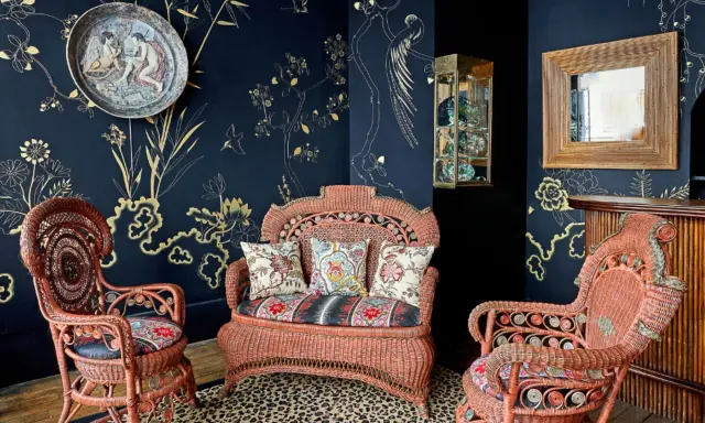Salon au style napoléon 3, avec des fauteuils et un canapé en rotin corail, sur un tapis léopard, devant un papier peint bleu nuit orné de motifs dorés.