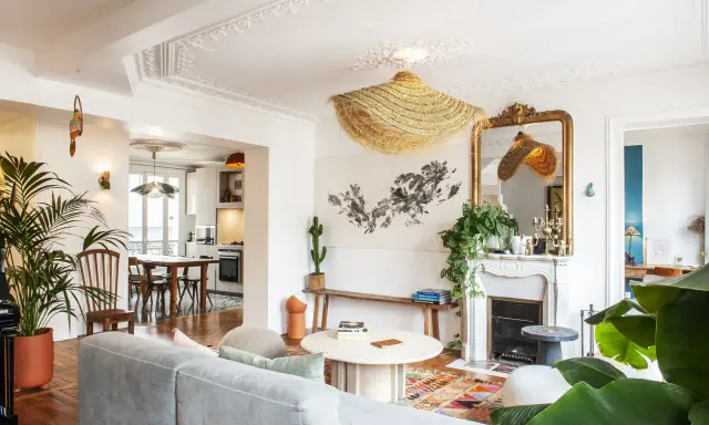 Salon au style classique revisité, avec un canapé gris, un tapis coloré, une cheminée ornée et des plantes luxuriantes.