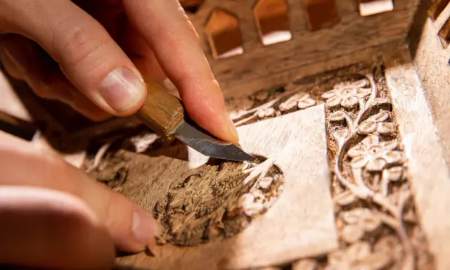 Main d'artisan sculptant un motif floral sur un meuble en bois, étape clé pour savoir comment nettoyer un meuble en marqueterie.