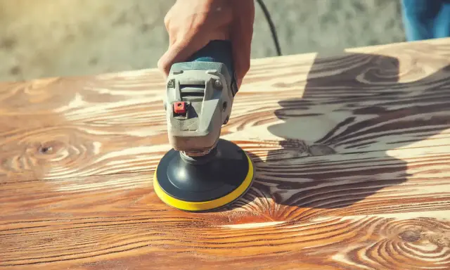 Main d'artisan utilisant une ponceuse pour enlever lasure sans poncer le bois. Le grain du bois est bien visible.