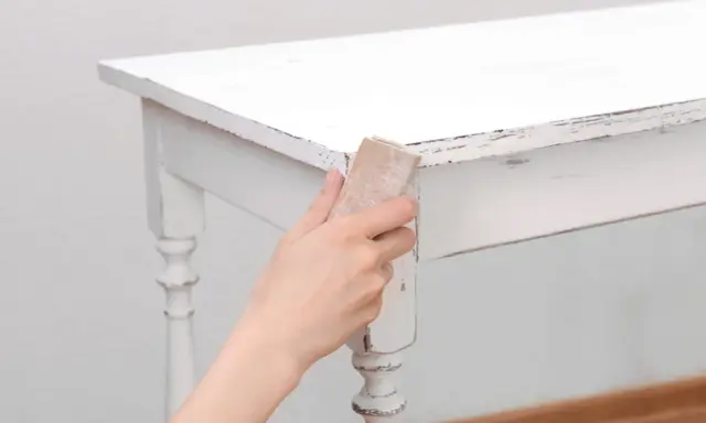Main tenant, comment décaper un meuble peint. Une main utilise du papier de verre pour poncer le bord d'une table blanche écaillée.
