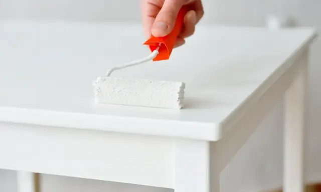 Main blanche d'un meuble laqué, un rouleau de peinture blanc est appliqué.