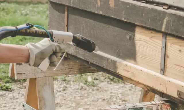 Main tenant en décapeur thermique pour enlever peinture sur bois. Le bois brut apparaît sous la couche sombre.