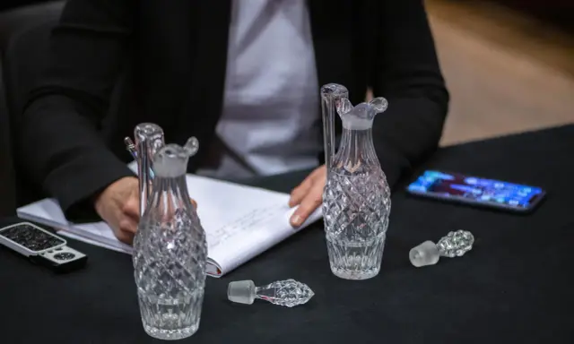 Une personne écrit sur un carnet, entourée de flacons en cristal. Une folle enchère se prépare pour ces objets précieux.