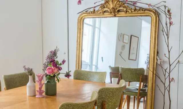 Un grand miroir ancien doré, orné de branches fleuries, reflète une salle à manger avec des chaises vertes. Quelle couleur peindre un miroir ancien ? L'or reste un classique intemporel.