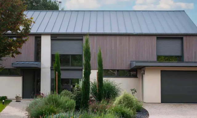 Façade moderne avec volet roulant maison ancienne, bois, pierre et jardin luxuriant.