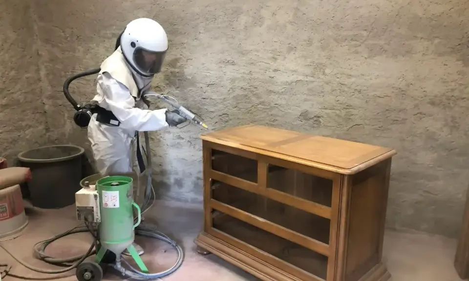 Personne en combinaison intégrale et casque effectuant un décapage bois sur une commode ancienne avec une machine de sablage.