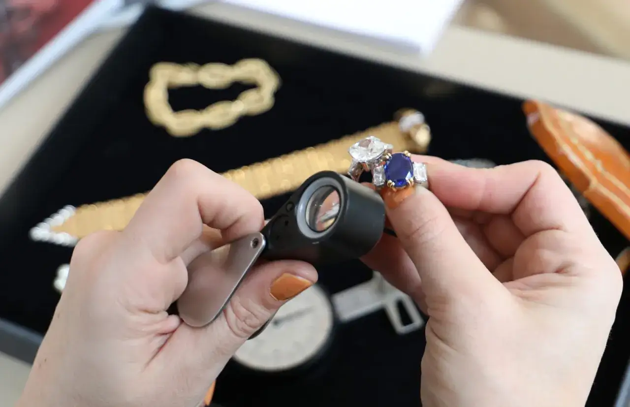 Main d'un expert en bijoux examinant une bague saphir et diamants avec une loupe.
