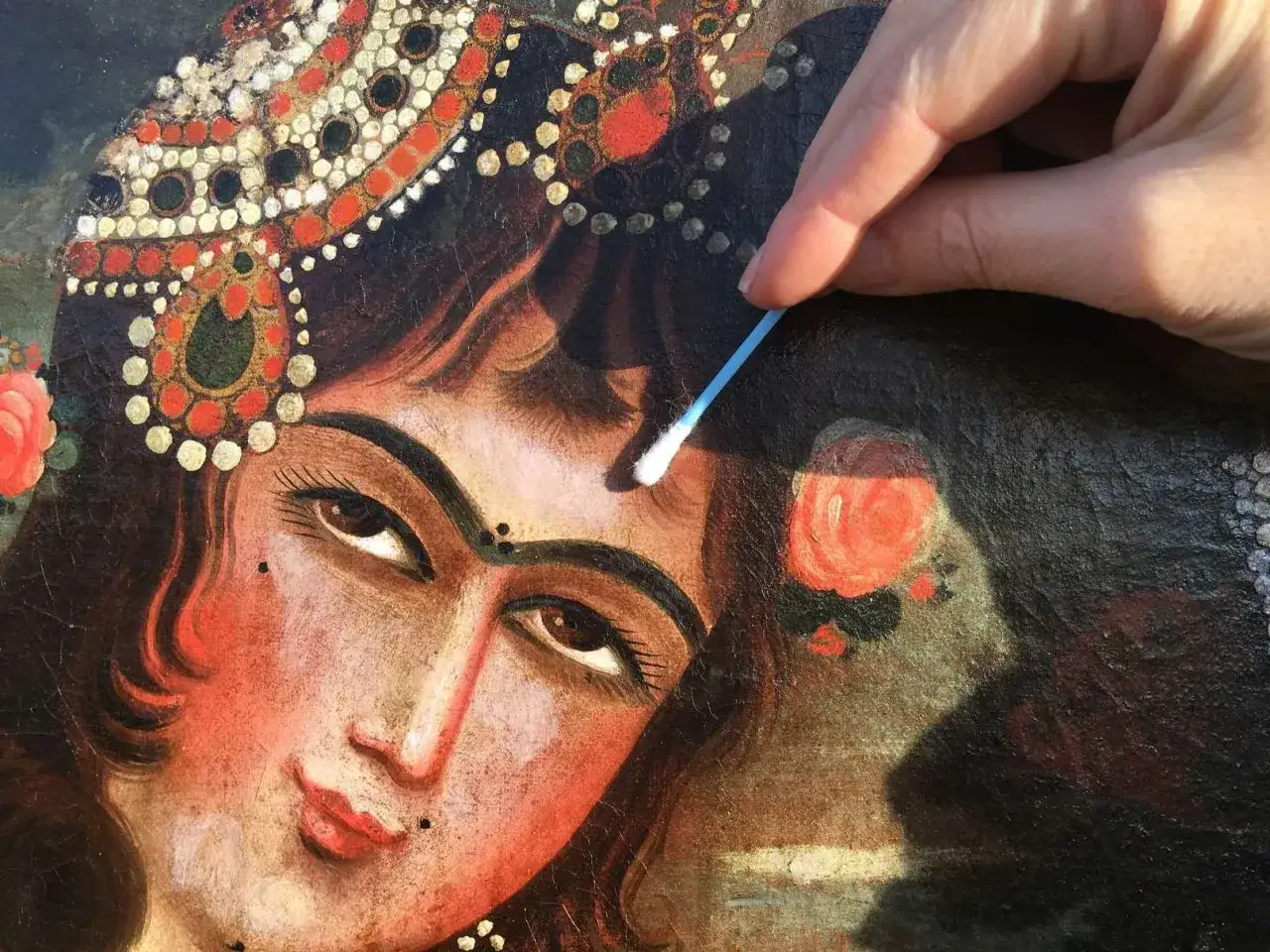 Main d'un restaurateur nettoyant d&eacute;licatement un tableau ancien avec un coton-tige, r&eacute;v&eacute;lant la beaut&eacute; d'un visage orn&eacute;.