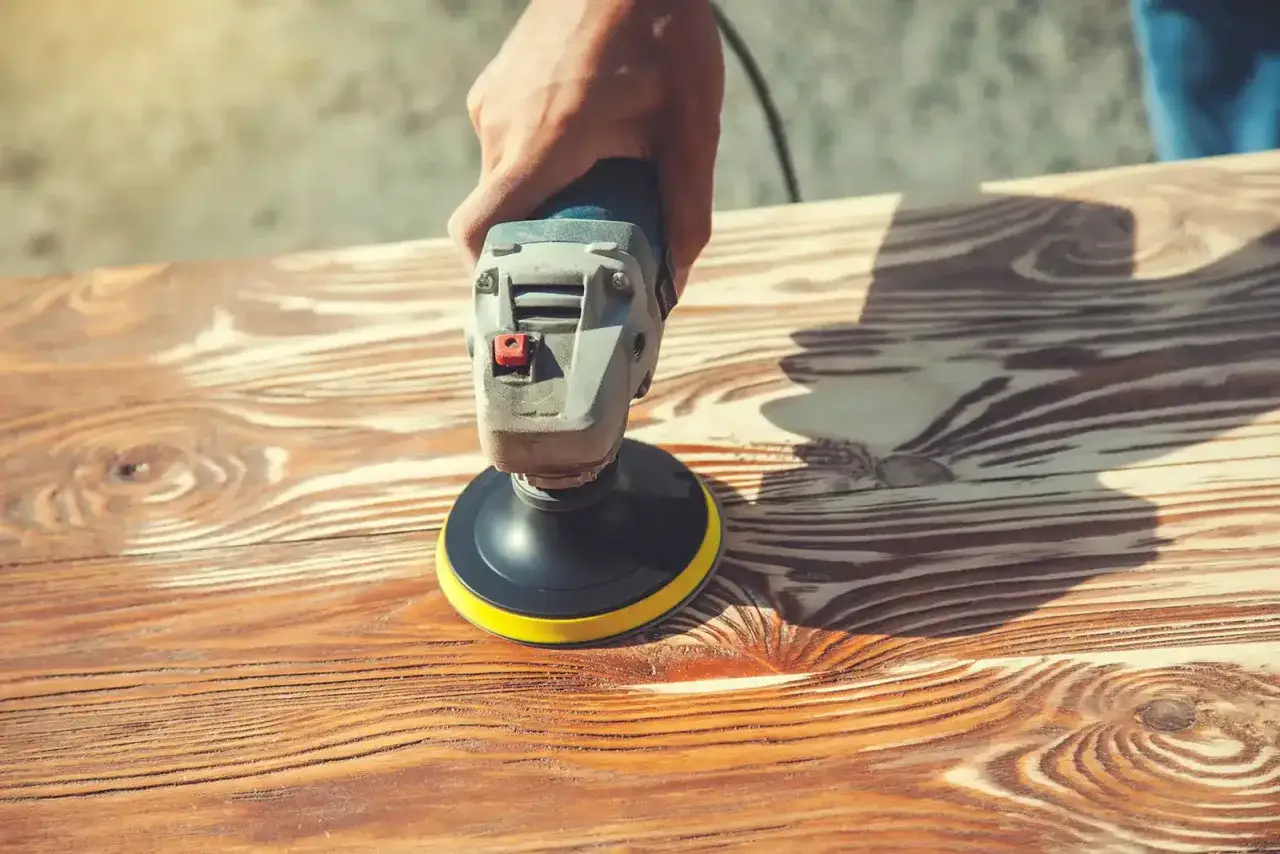 Main d'artisan utilisant une ponceuse pour enlever lasure sans poncer le bois. Le grain du bois est bien visible.