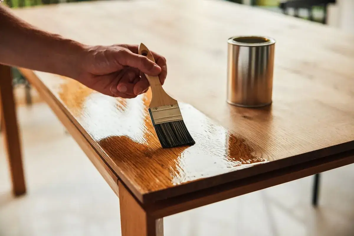 Main d'artisan appliquant une couche de vernis sur une table en bois, lui redonnant son &eacute;clat.