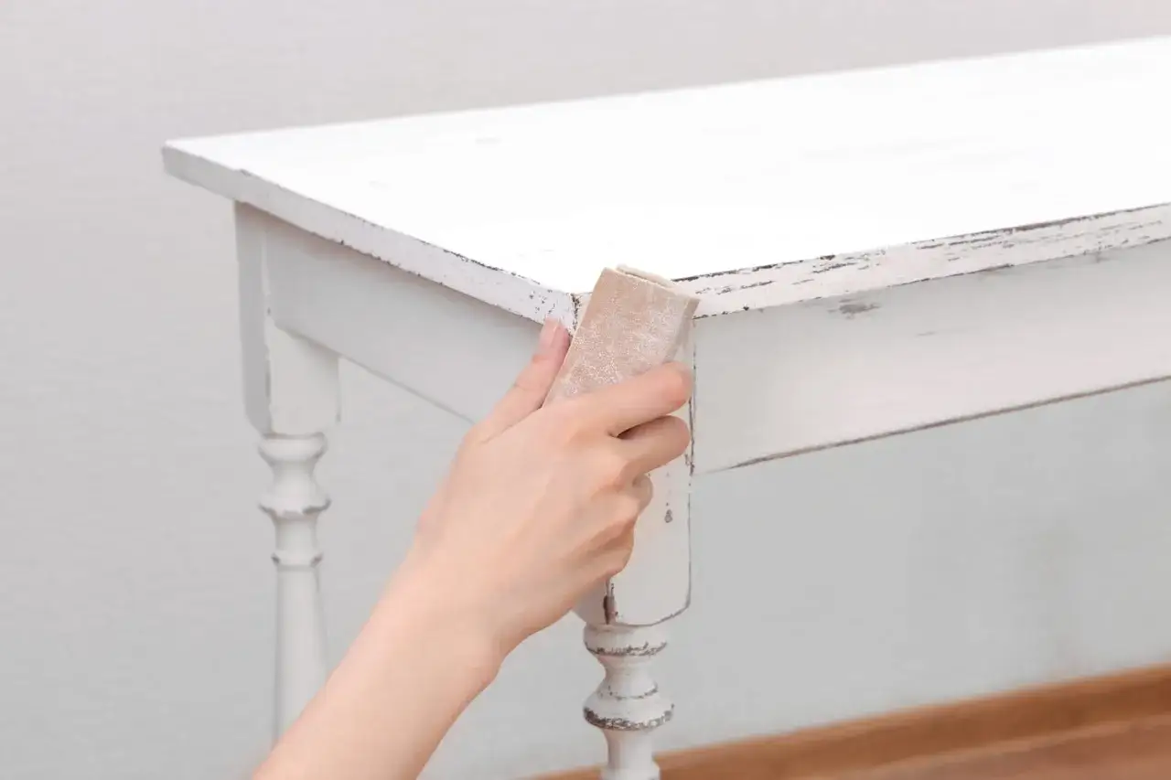 Main tenant, comment décaper un meuble peint. Une main utilise du papier de verre pour poncer le bord d'une table blanche écaillée.