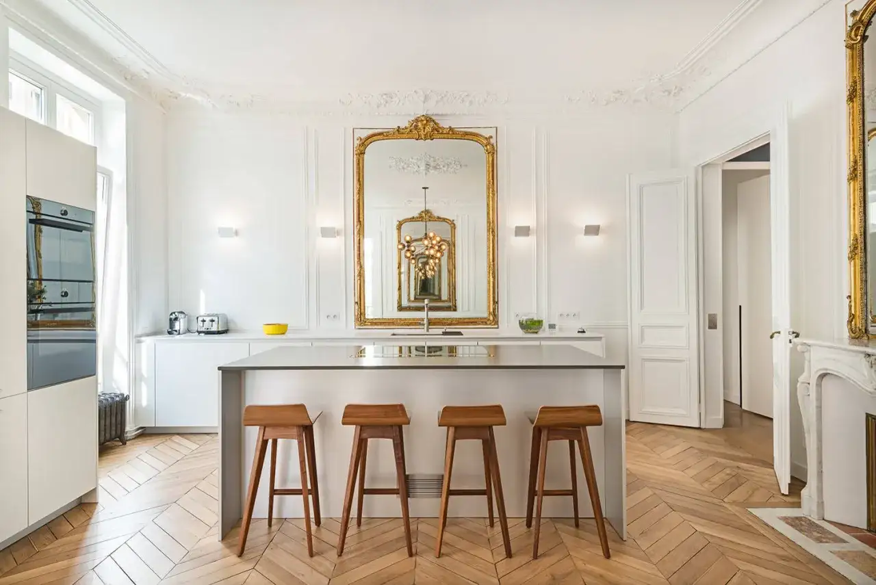 Cuisine lumineuse au style classique, avec un grand miroir dor&eacute;, un &icirc;lot central et des tabourets en bois sur un parquet en chevrons.