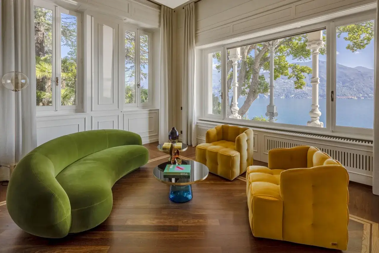 Salon avec canapé vert courbé et fauteuils jaunes, dans un style art déco, donnant sur un lac.